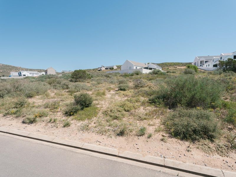 A vacant residential plot of land featuring natural coastal vegetation, accessible from a paved road with established concrete curbing.