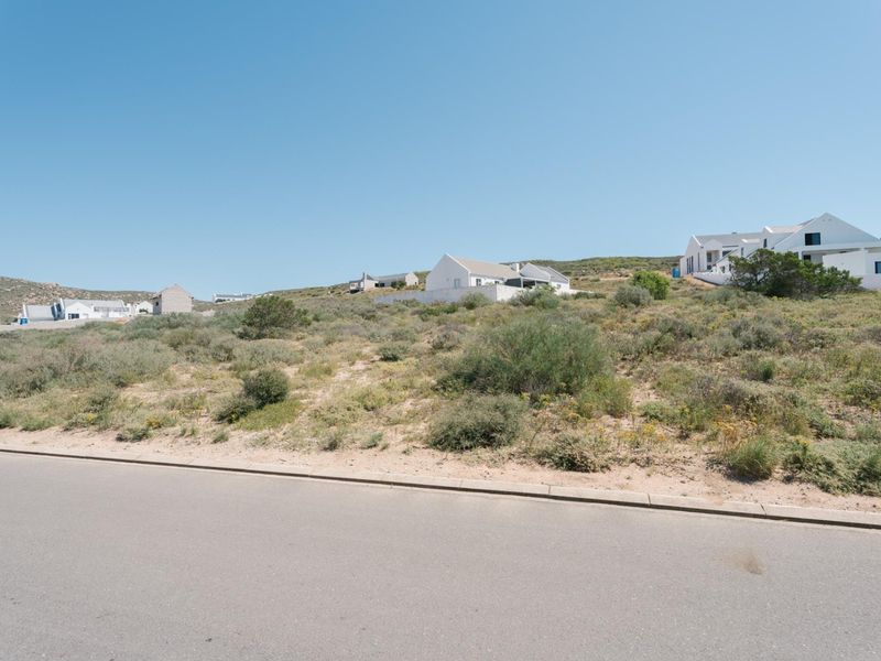 A street-level view of a vacant residential plot featuring natural coastal vegetation, situated in a developed neighborhood with existing homes nearby.