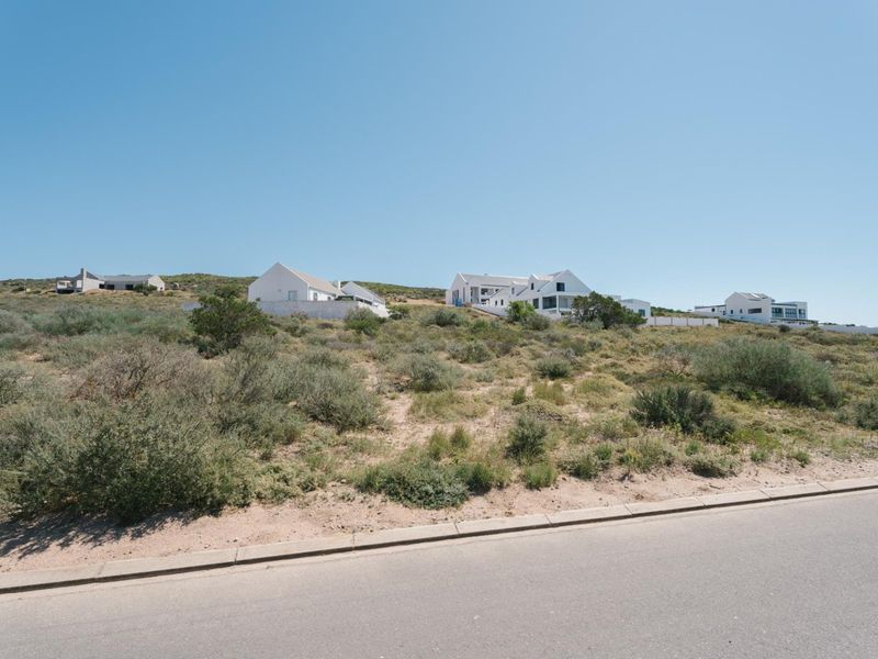 A view of a vacant residential plot from the adjacent paved road, featuring natural coastal vegetation and surrounding residential properties.