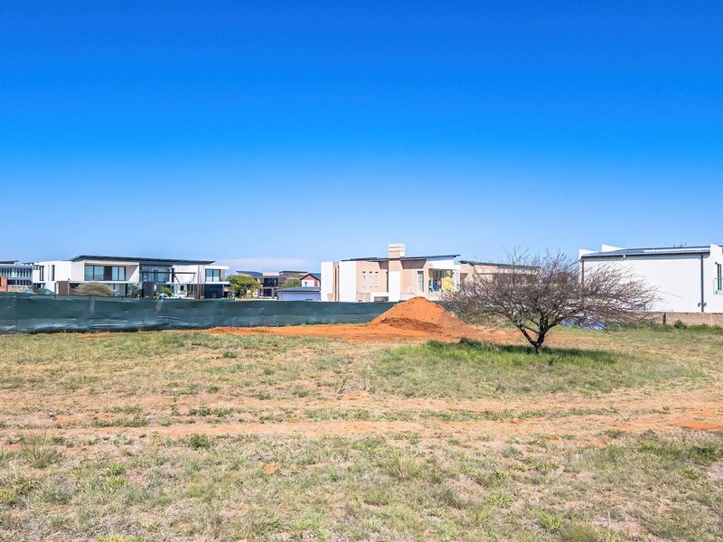 A view of a vacant residential plot featuring natural grass, a small tree, and perimeter fencing, situated within a developed residential estate.