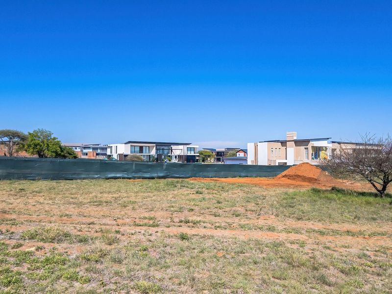 A vacant residential plot of land featuring natural grass coverage and perimeter fencing, situated within a developed residential estate.