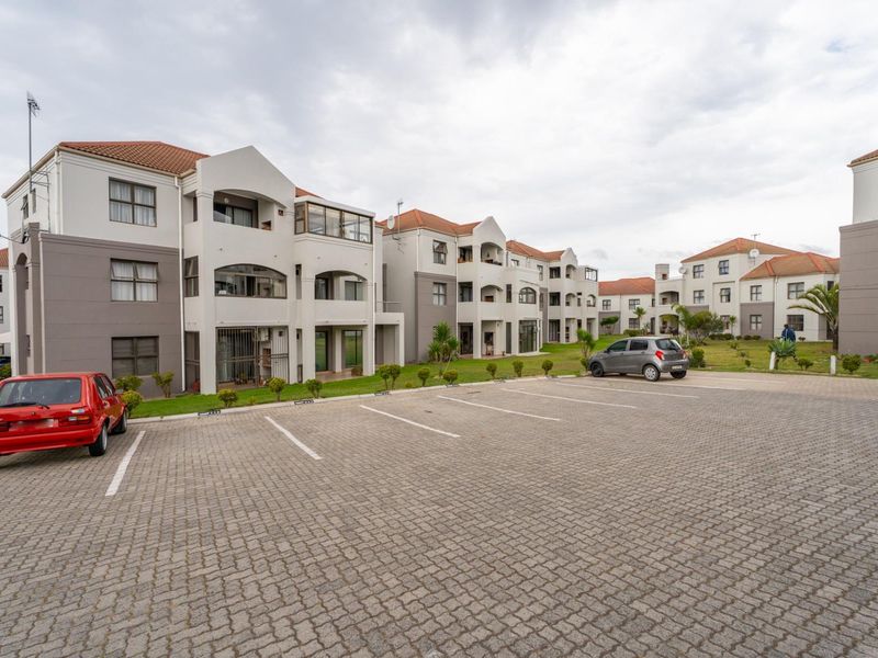 Exterior view of a multi-story apartment complex featuring paved parking, landscaped grounds, and private balconies with terracotta roof tiles.
