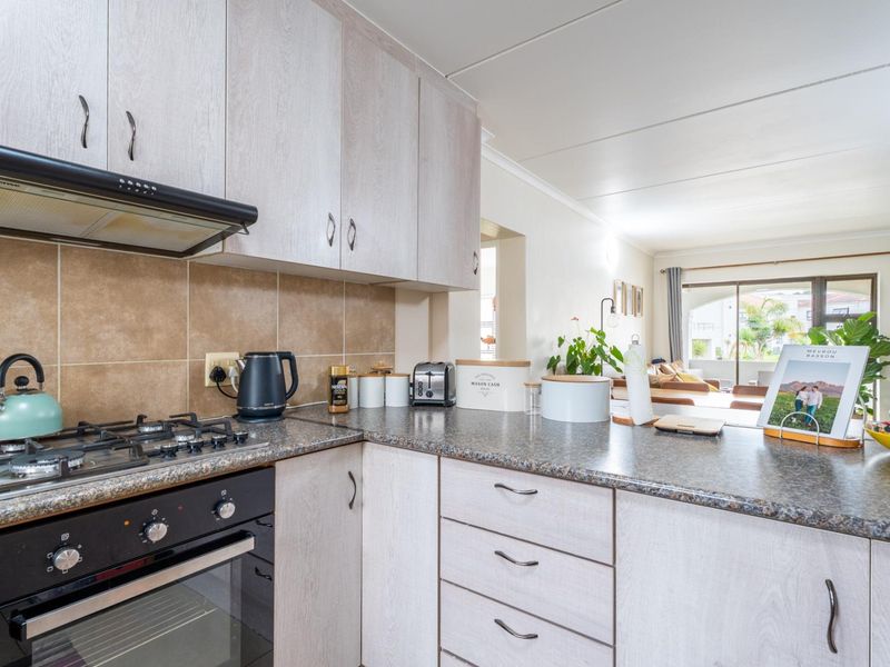 Open-plan kitchen featuring light wood-finish cabinetry, granite countertops, a gas hob, and an integrated oven, flowing into a bright living area.