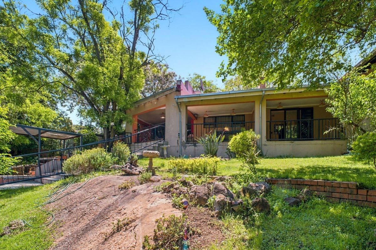 Rear view of the property featuring a covered patio with wrought iron railings, overlooking a landscaped garden with natural rock features and brick retaining walls.