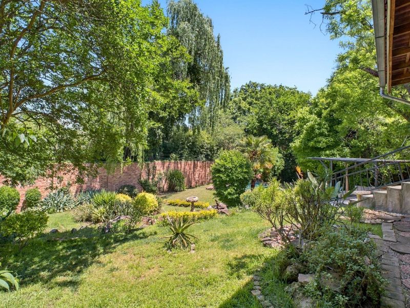 A lush, well-established garden featuring mature trees, landscaped shrubbery, a brick boundary wall, and a stone pathway leading to the rear of the property.