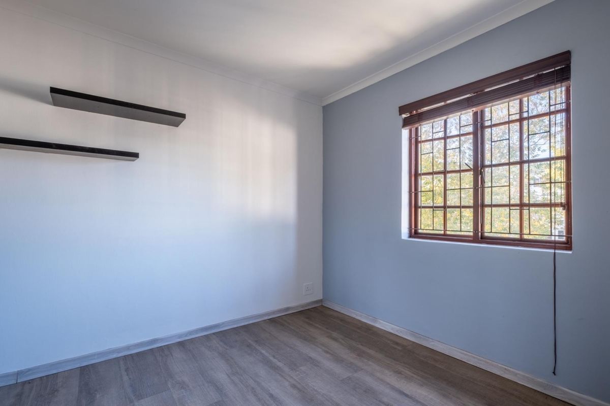 A bright, empty bedroom featuring modern laminate flooring, a window with security bars, and built-in floating shelves.