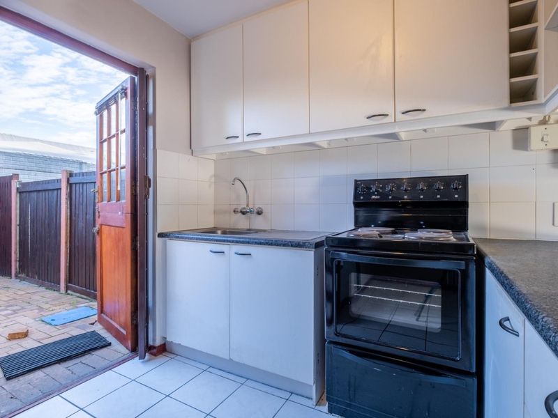 A compact kitchen featuring white cabinetry, laminate countertops, a tiled backsplash, and a freestanding electric stove, with direct access to an outdoor paved area.