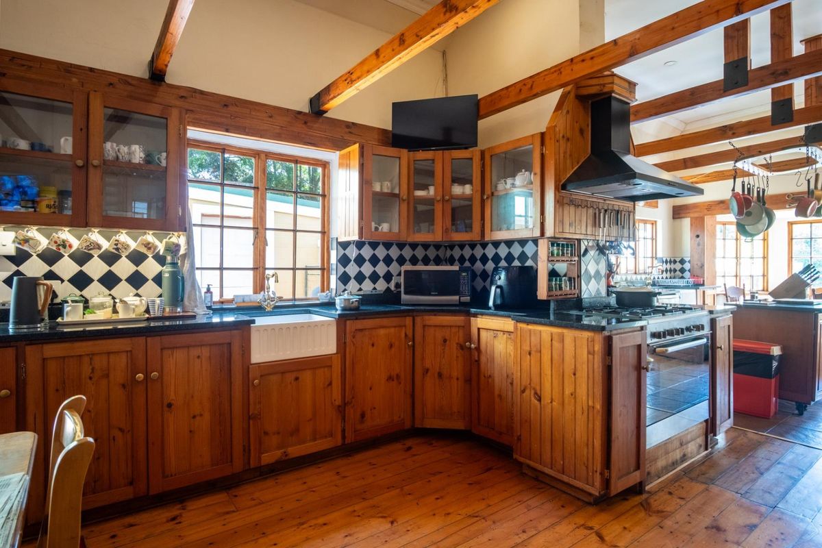 A rustic-style kitchen featuring exposed timber beams, warm wood cabinetry, black granite countertops, a farmhouse sink, and a classic checkered backsplash.