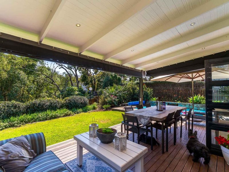 A spacious covered timber deck patio overlooking a lush garden and swimming pool, featuring exposed beam ceilings and a retractable awning for shade.
