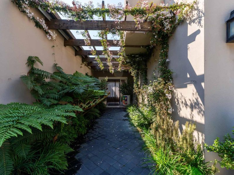 A charming, landscaped entrance walkway featuring a timber pergola, lush greenery, and paved flooring leading to the front door.