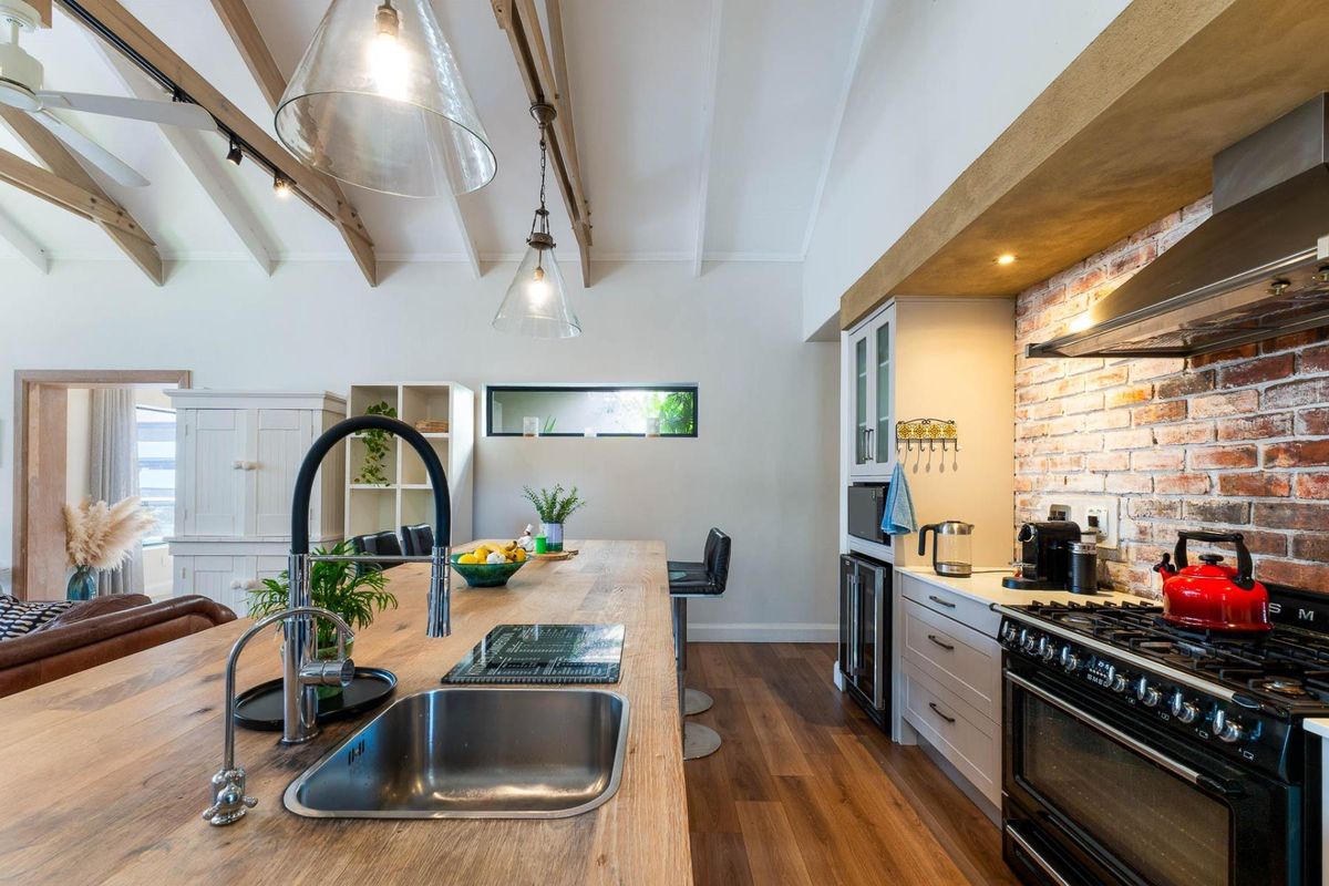 A modern kitchen featuring a large timber island, exposed brick wall, gas range cooker, and vaulted ceilings with exposed timber beams.