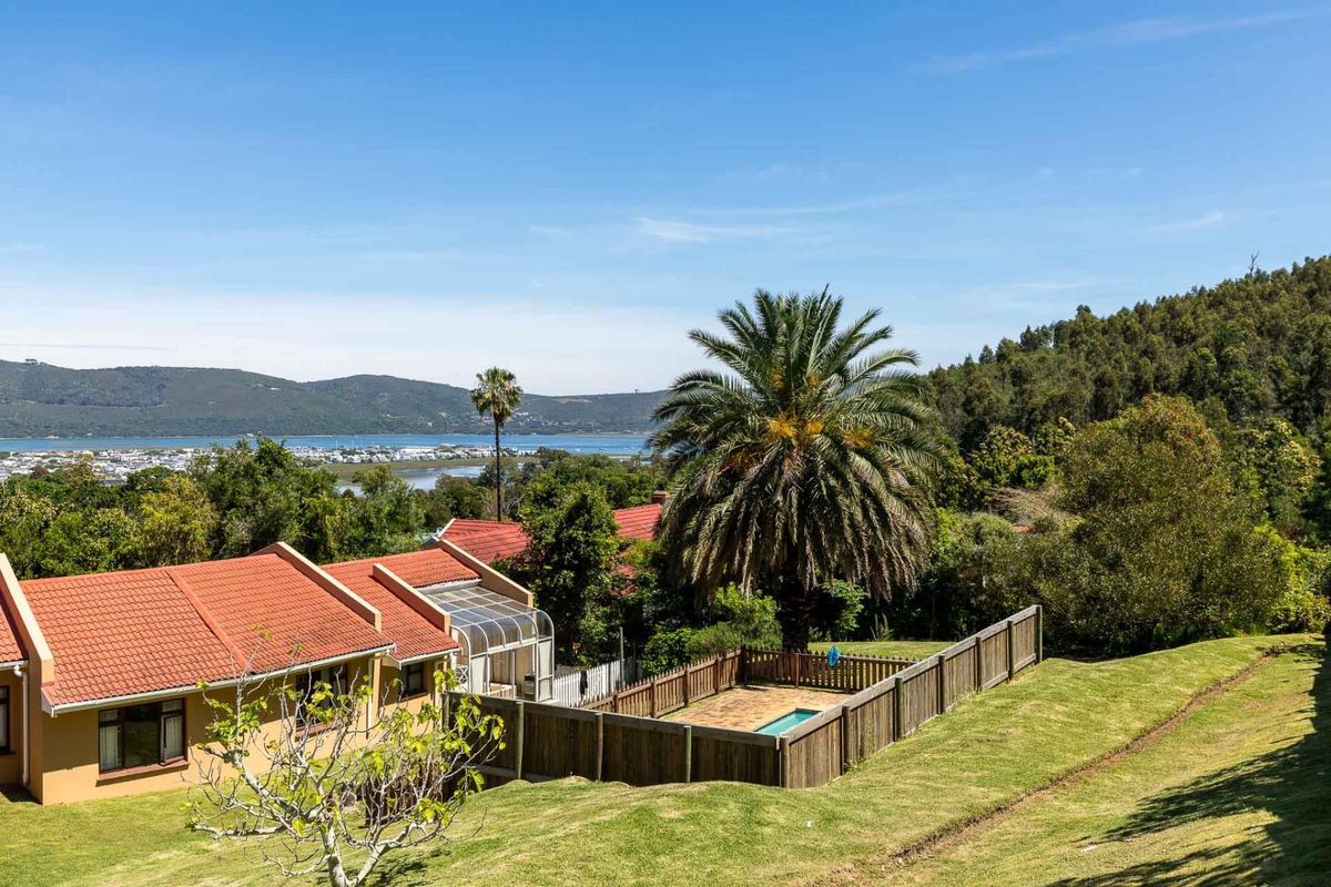Elevated view of the property featuring a swimming pool with timber fencing, tiled roof structure, and expansive garden overlooking the lagoon and mountains.