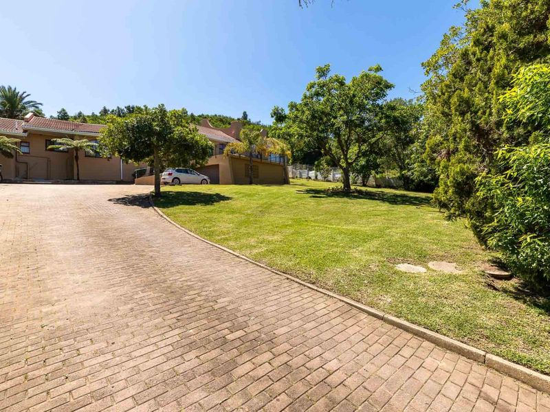 A spacious front exterior view featuring a paved driveway leading to the house, surrounded by a well-maintained garden and mature trees.
