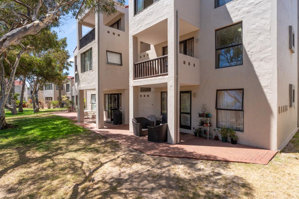 Rear view of a multi-story apartment building featuring private balconies, ground-floor paved patios, and landscaped garden areas.