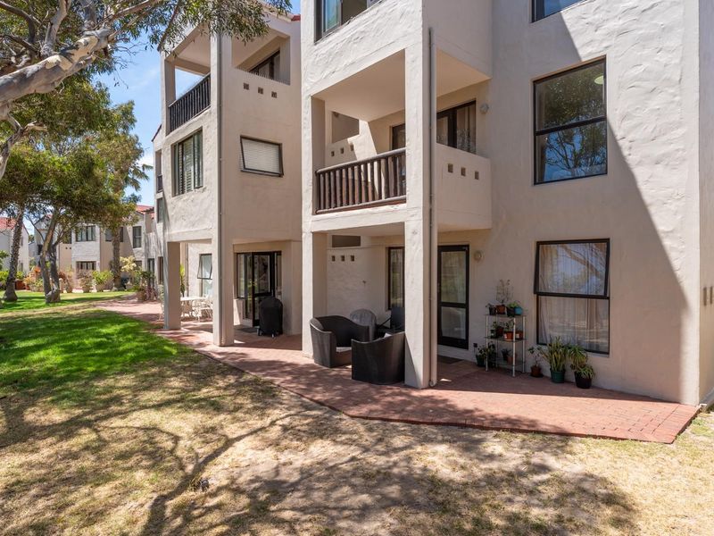 Rear view of a multi-story apartment building featuring private balconies, ground-floor paved patios, and landscaped garden areas.