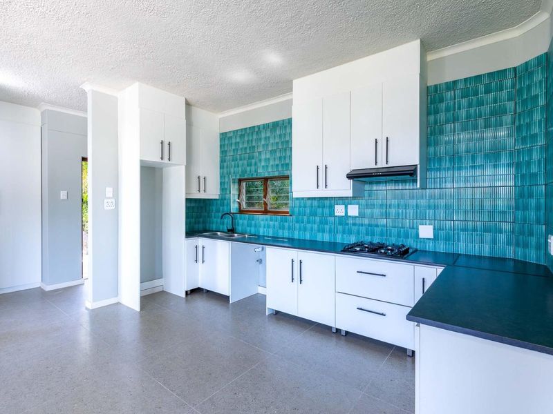 A modern, bright kitchen featuring white cabinetry, dark stone countertops, and a striking teal tiled splashback with a built-in gas hob and extractor.