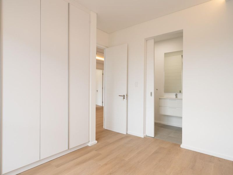 A bright, modern bedroom featuring light laminate flooring, ample built-in cupboard space, and direct access to an en-suite bathroom with a vanity.