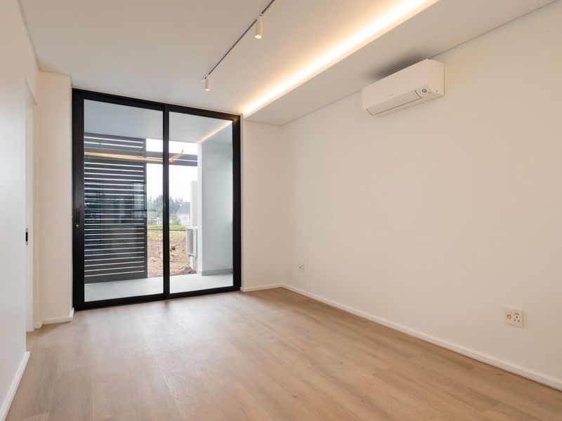 A bright, modern bedroom featuring light wood-look flooring, a split-system air conditioner, recessed lighting, and a sliding glass door leading to a private balcony.