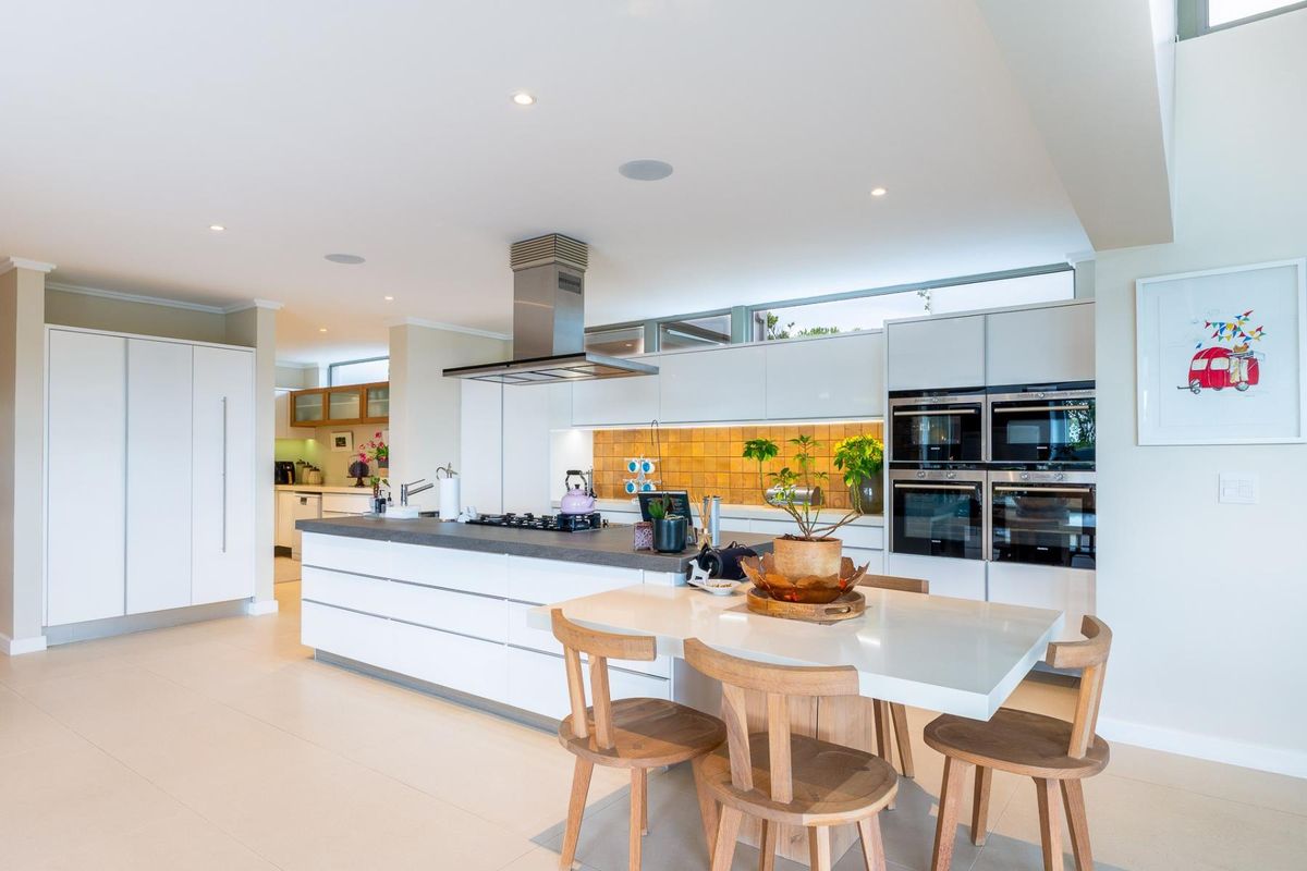 Modern open-plan kitchen featuring a large island, built-in ovens, gas hob, extractor fan, and sleek white cabinetry with a warm tiled splashback.