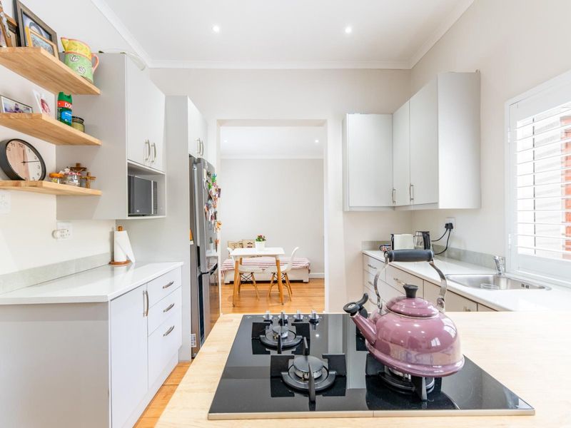 Modern kitchen featuring light grey cabinetry, stone countertops, a gas cooktop, and floating timber shelving, with an adjacent dining area visible.