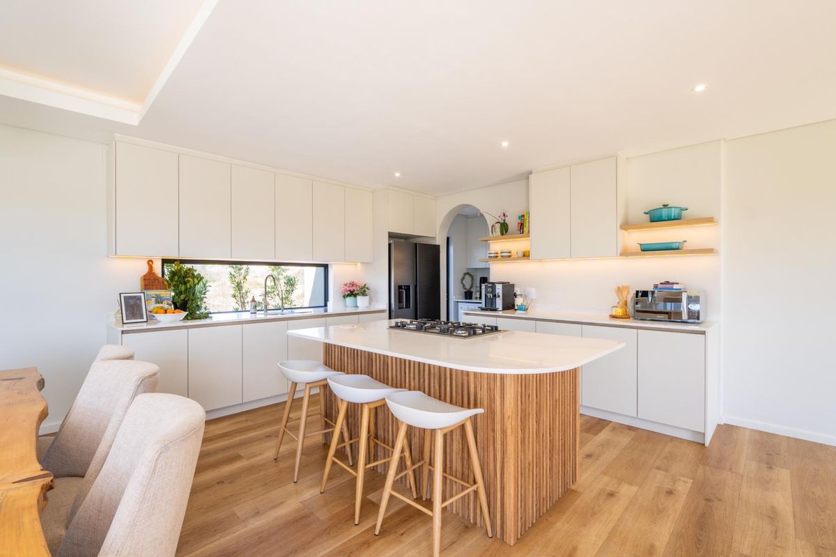 Modern kitchen featuring a central island with gas cooktop, sleek handleless cabinetry, floating shelves, and warm hardwood flooring with integrated lighting.