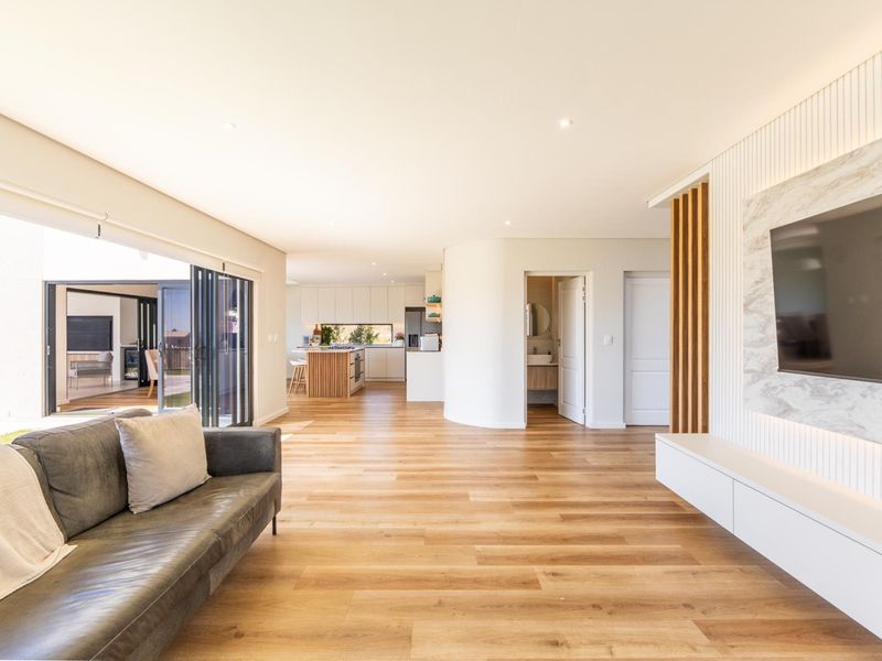 Spacious open-plan living area featuring timber flooring, recessed lighting, a modern marble media wall, and seamless indoor-outdoor flow through large stacking glass doors.
