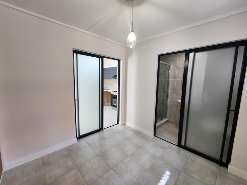 An open-plan living area featuring modern tiled flooring, sleek black-framed sliding doors, and contemporary pendant lighting, providing access to the kitchen and bathroom.