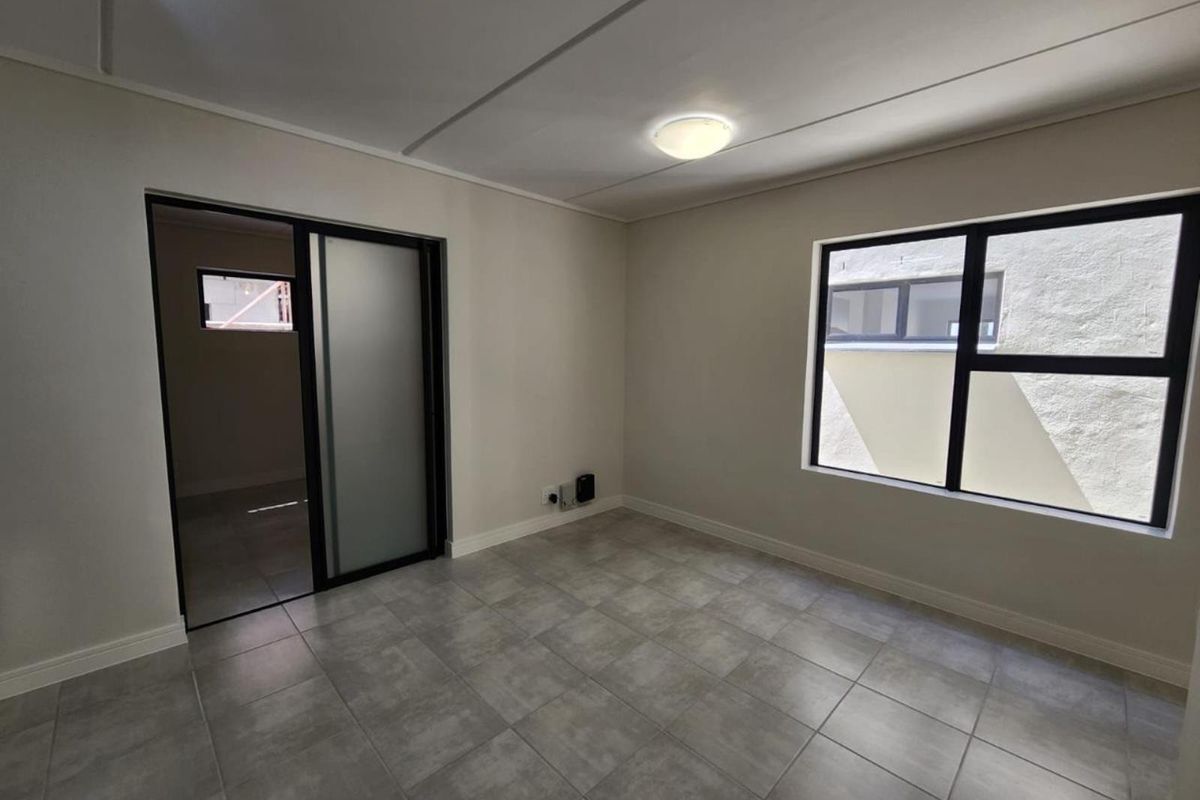 A clean, empty bedroom featuring neutral tiled flooring, modern aluminium window frames, and a sliding door leading to an adjacent space.