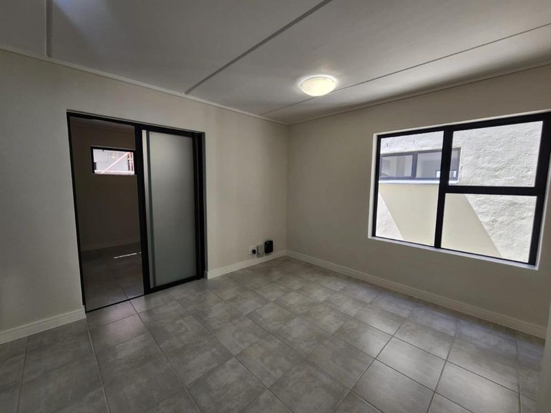 A clean, empty bedroom featuring neutral tiled flooring, modern aluminium window frames, and a sliding door leading to an adjacent space.