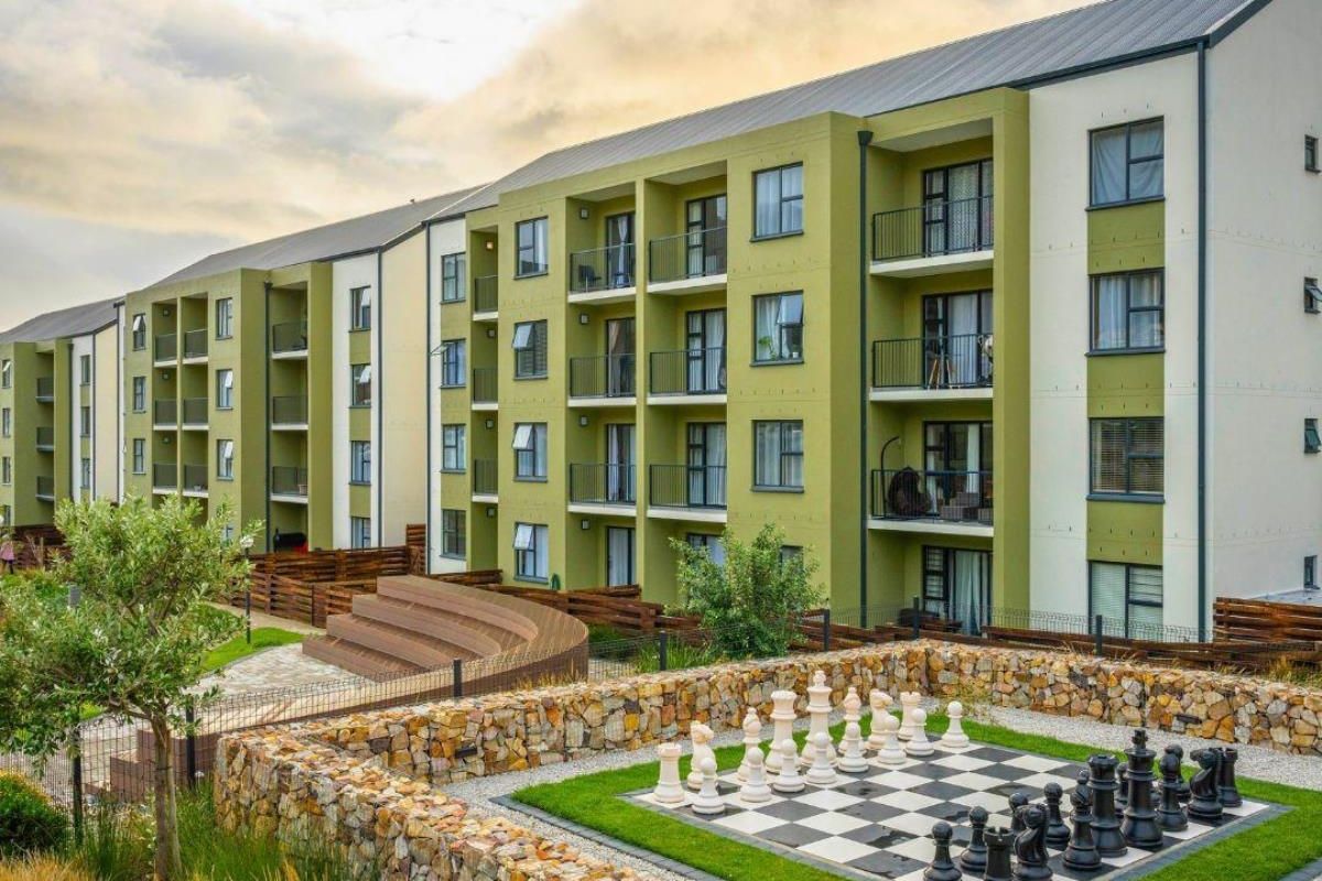 Exterior view of a modern apartment complex featuring private balconies, landscaped grounds, and a communal outdoor chess area with stone retaining walls.