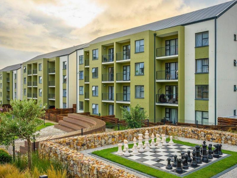 Exterior view of a modern apartment complex featuring private balconies, landscaped grounds, and a communal outdoor chess area with stone retaining walls.