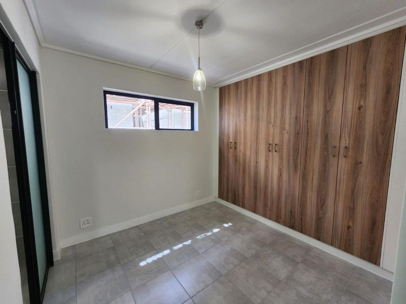 A clean, modern bedroom featuring neutral tiled flooring, ample built-in wooden cupboards, and a contemporary pendant light fixture.