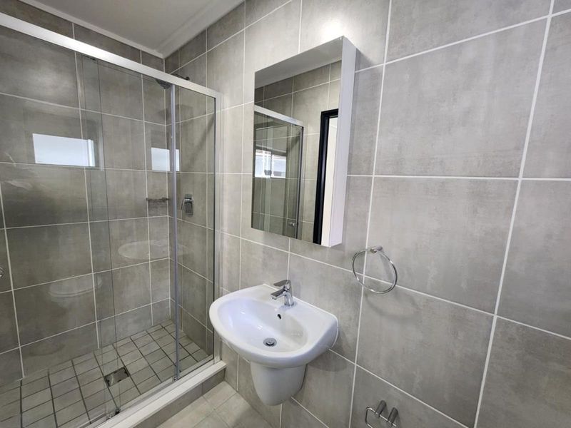 A modern, clean bathroom featuring a glass shower enclosure, pedestal washbasin, and neutral grey ceramic wall tiling.
