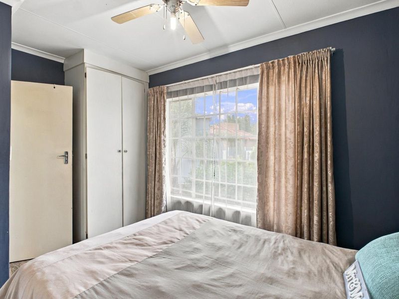 A bedroom featuring dark blue walls, a built-in wardrobe, and a ceiling fan, with natural light entering through a grid-pattern window.