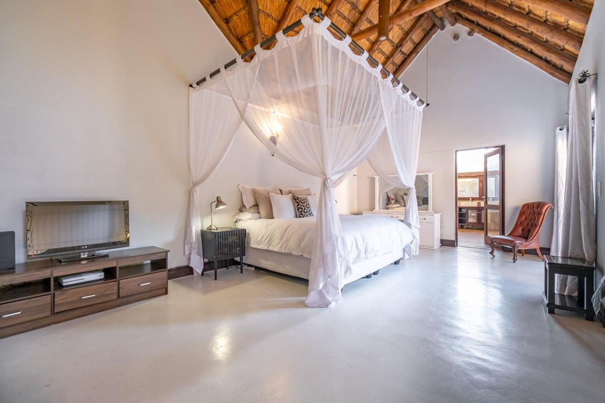 Spacious bedroom featuring high vaulted ceilings with exposed thatch and timber rafters, complemented by polished concrete flooring and direct access to an en-suite bathroom.