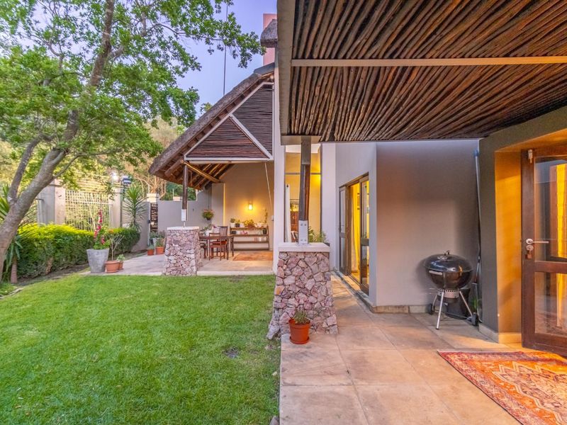 A spacious covered patio featuring a thatched roof and stone-clad pillars, overlooking a well-maintained garden area with direct access to the interior living space.