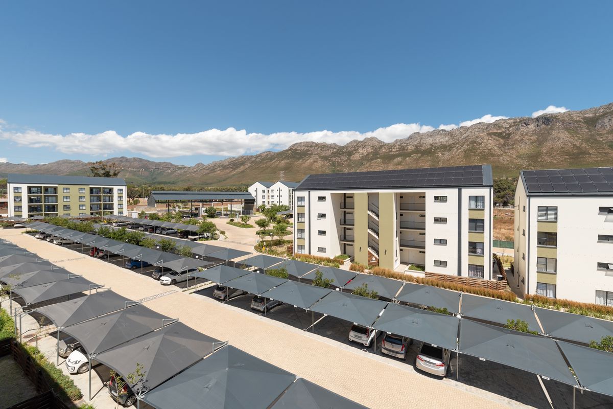 Aerial view of a modern apartment complex featuring multiple residential buildings with solar panels and extensive covered parking facilities set against a mountain backdrop.