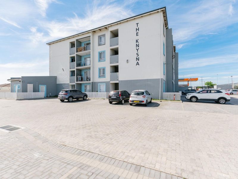 Exterior view of The Knysna apartment complex featuring a paved parking area, multi-story building facade, and private balconies.
