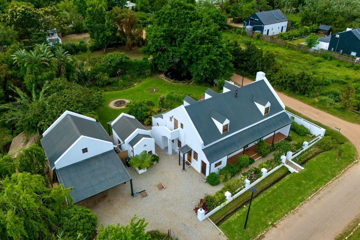 Aerial view of a well-maintained property featuring a main residence with a metal roof, dormer windows, a gravel driveway, and a landscaped garden.