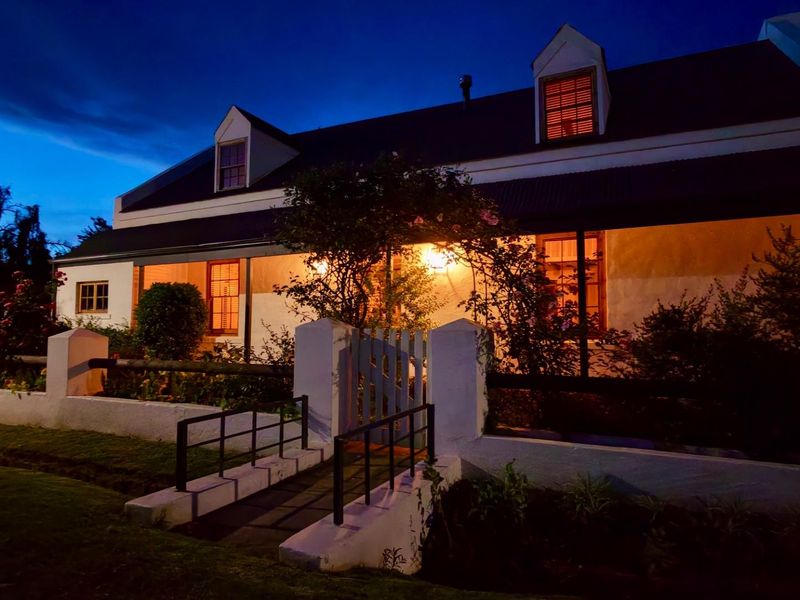 Charming cottage-style home exterior at dusk, featuring a covered veranda, dormer windows, and a landscaped front garden with a pedestrian footbridge.