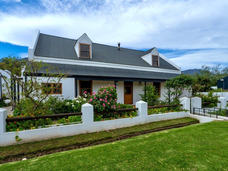 Charming Cape Dutch style cottage featuring a corrugated metal roof, dormer windows, and a welcoming front veranda set behind a landscaped garden and boundary wall.