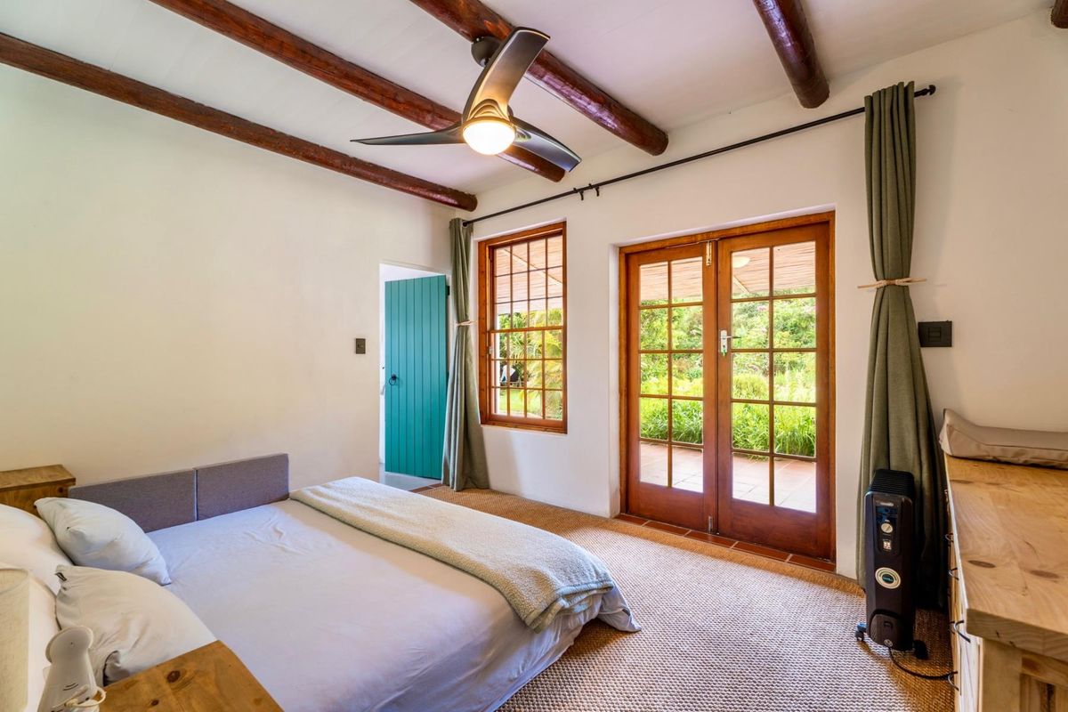 A bright bedroom featuring exposed timber ceiling beams, a ceiling fan, and timber-framed french doors providing direct access to the garden.