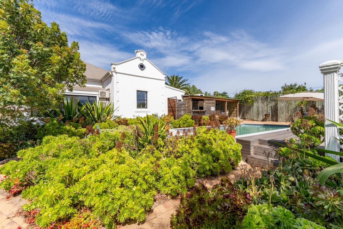 A well-maintained rear garden featuring a swimming pool, timber decking, and a covered patio area with a built-in braai, set against a historic home.