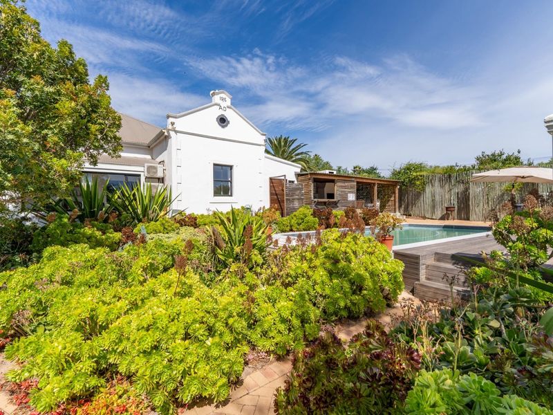 A well-maintained rear garden featuring a swimming pool, timber decking, and a covered patio area with a built-in braai, set against a historic home.