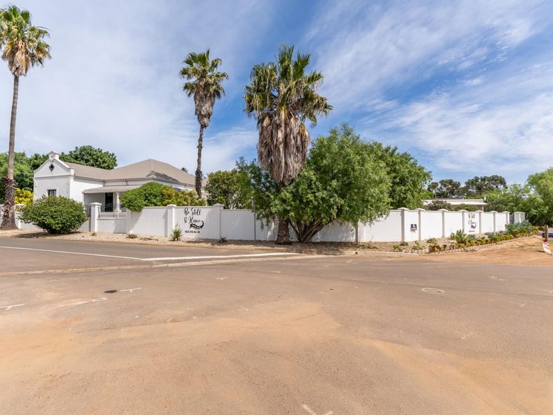 A well-maintained property exterior featuring a white perimeter wall, gabled roof architecture, and established palm trees, viewed from the street corner.