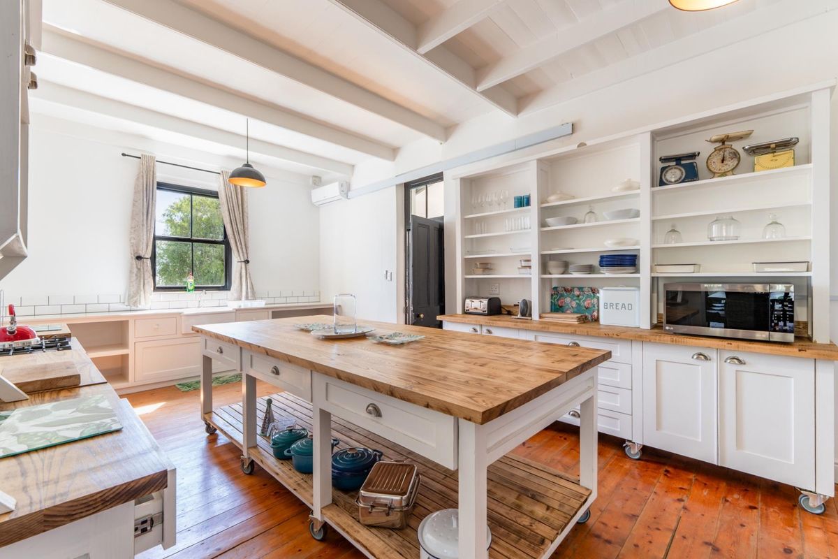 A bright, farmhouse-style kitchen featuring a large timber-topped island, butler sink, open shelving, and exposed white-painted ceiling beams over hardwood floors.