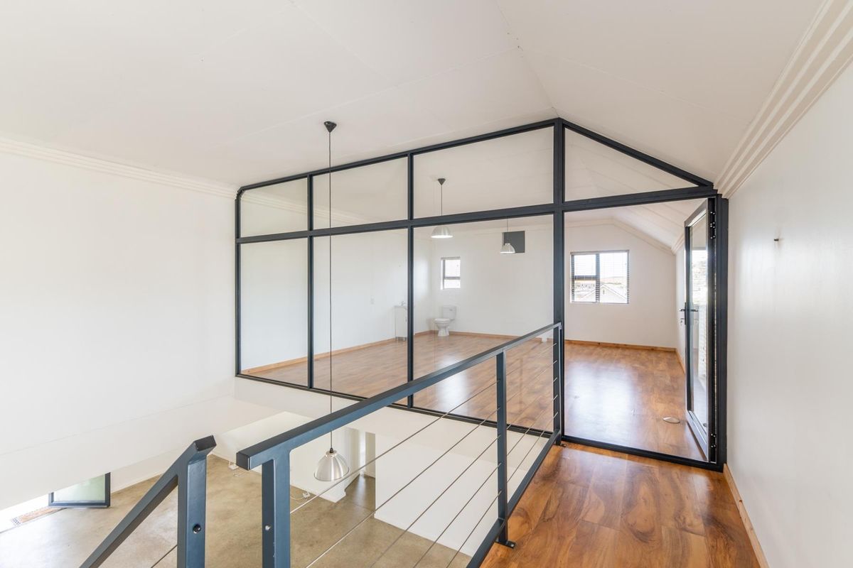 A bright, open-plan bedroom area featuring a modern glass partition, vaulted ceilings, and laminate flooring, overlooking the lower level via a metal staircase railing.