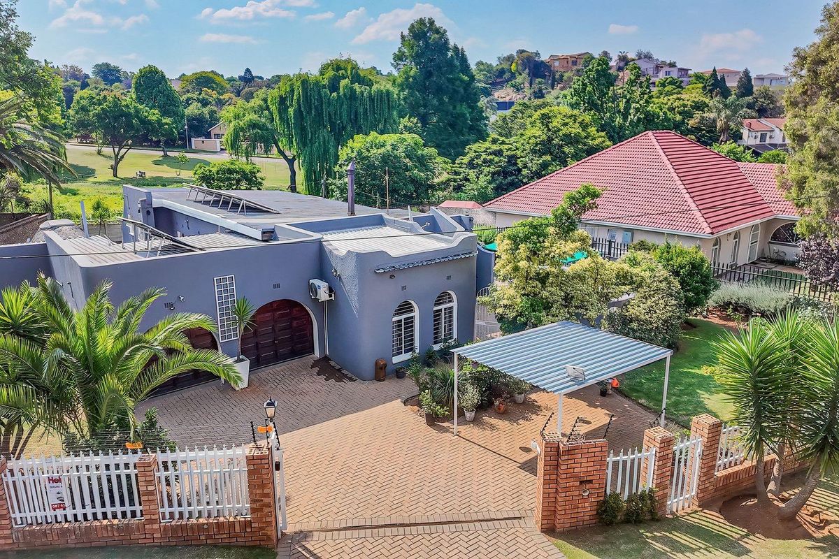 Aerial view of a residential property featuring a paved driveway, carport, solar panels, and established garden with brick boundary wall and electric fencing.