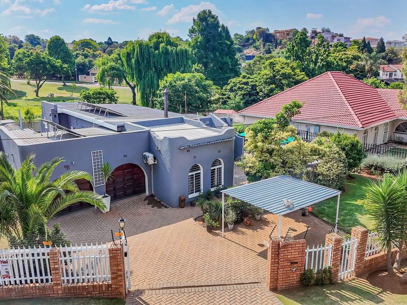 Aerial view of a residential property featuring a paved driveway, carport, solar panels, and established garden with brick boundary wall and electric fencing.