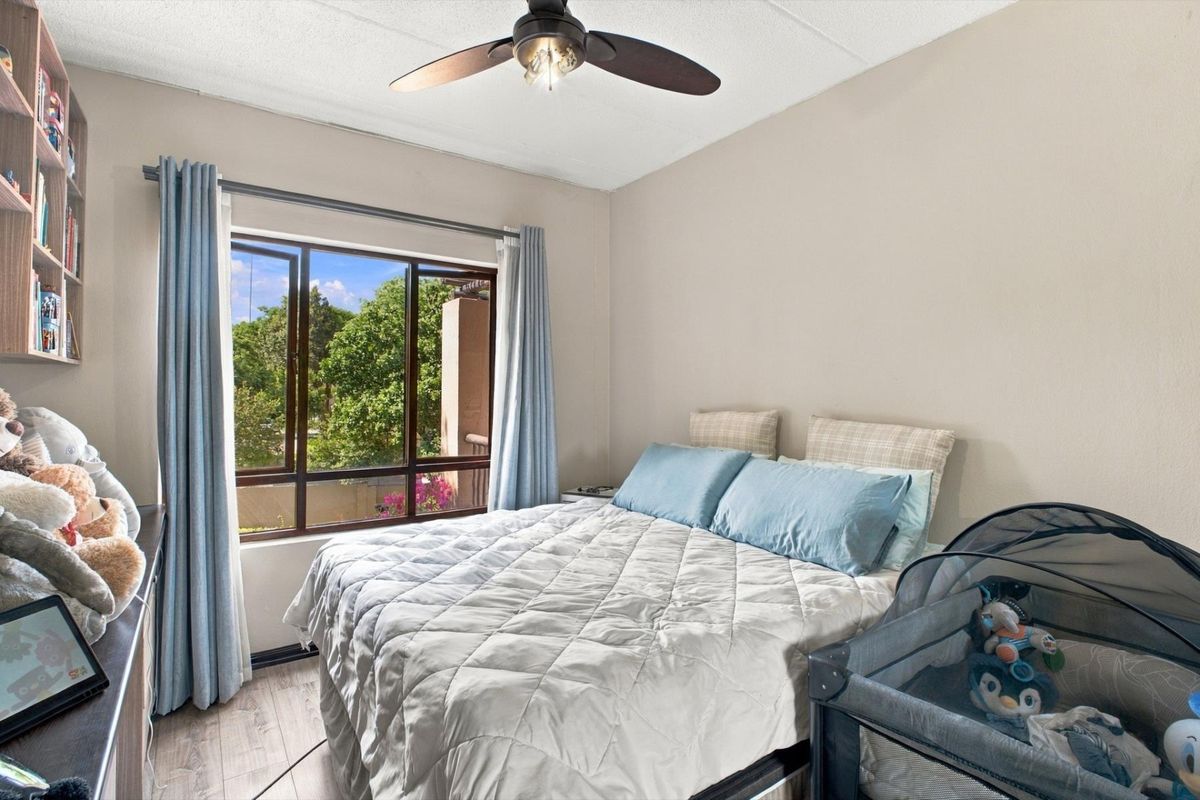 A well-lit bedroom featuring laminate flooring, a ceiling fan, and built-in shelving, with a large window providing natural light and exterior views.